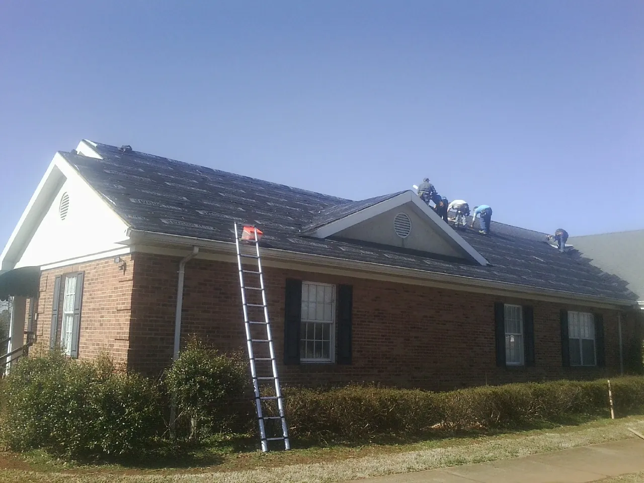 Crew tearing shingles off a brick ranch roof with a ladder set — Emergency Roof Repair work by our crew in Reasnor