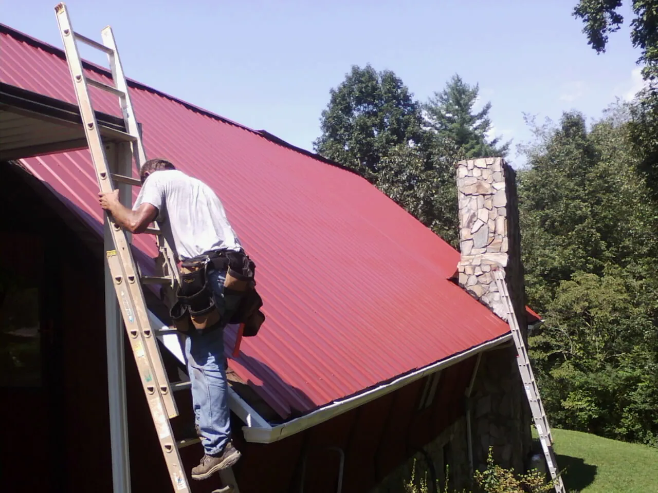 Roofer on a ladder beside a red metal roof and stone chimney — Soffit & Fascia Repair by Roofing Brothers in Reasnor